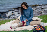 A woman sits on a cream blanket on grass by the water, wearing a blue denim dress and patterned knee-high socks. A small fire pit with glowing embers sits nearby, with rocks and water in the background.