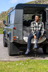 A man sitting on the open rear hatch of a dark green Land Rover on a gravel road outdoors, wearing a green and white plaid flannel shirt, blue jeans, and fairisle socks.
