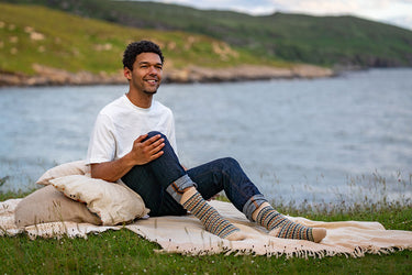 A man sitting on a cream blanket on a grassy shore by the water, wearing a white t-shirt, dark jeans, and patterned knit socks. Green hills and a body of water are in the background.