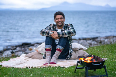 A man sits on a blanket by the ocean with a small fire burning in a black grill. He is wearing a green and black plaid flannel shirt, blue jeans, and patterned knit socks.