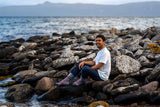 A man sitting on large rocks by the water, wearing a white t-shirt, dark jeans, and striped socks, with a rocky shoreline and distant water in the background.