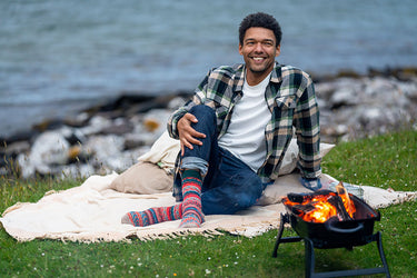 A man sitting on a blanket by the water, wearing a plaid shirt, jeans, and colorful striped socks, with a small portable fire pit on the grass in front of him.