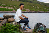 A man sitting on a rock by the water wearing a white t-shirt, dark jeans, and colorful fairisle socks, with a lobster trap on the ground nearby and a green hill in the background.