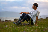 A man sits on a grassy hill in a field of wildflowers, looking off into the distance. He is wearing a white t-shirt, dark jeans, and colorful patterned socks, with a cloudy sky and distant hills in the background.