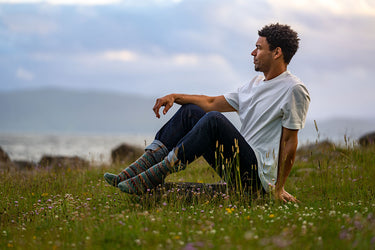 A man sits on a grassy hill in a field of wildflowers, looking off into the distance. He is wearing a white t-shirt, dark jeans, and colorful patterned socks, with a cloudy sky and distant hills in the background.