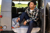 A man sitting on the open rear door of a gray vehicle, wearing a green and black plaid flannel shirt, dark blue jeans, and colorful striped socks.