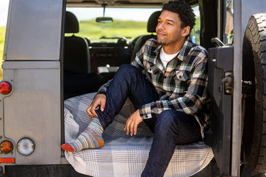 A man sitting on the open rear door of a gray vehicle, wearing a green and black plaid flannel shirt, dark blue jeans, and colorful striped socks.