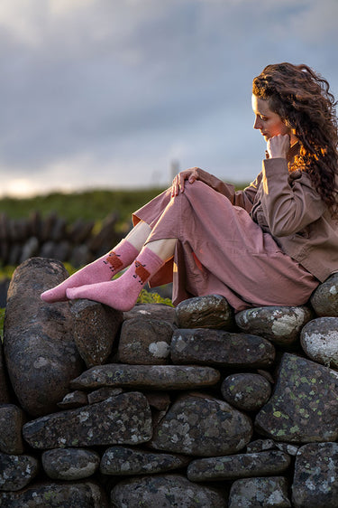 A woman sits on a stone wall outdoors, wearing a dusty pink outfit and pink socks with a brown cow print. The background shows a cloudy sky and green grassy hills.