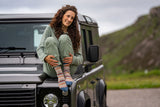 A woman with curly brown hair sits on the hood of a dark gray SUV outdoors, wearing sage green loungewear and colorful knit socks, with a blurred mountainous landscape in the background.